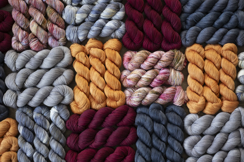 Hand-dyed yarn and dried flowers laid out in a grid in colors of grey, deep red, gold, slate, pink and more