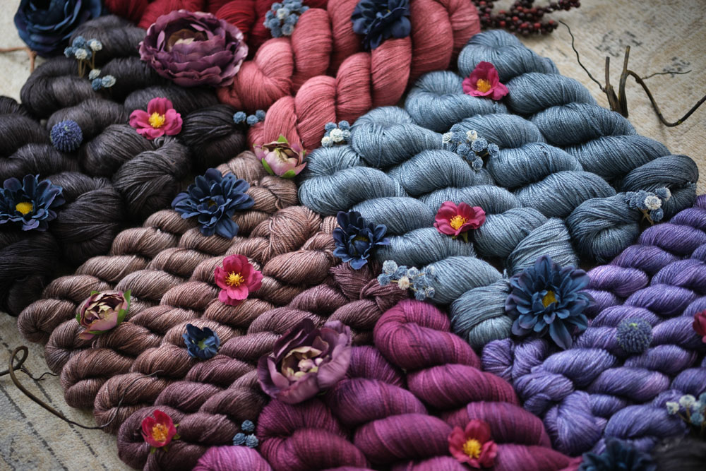 A pile of hand-dyed yarn and dried flowers in shades of raspberry, smokey teal, periwinkle, cocoa black, scarlet red, copper and umber and more