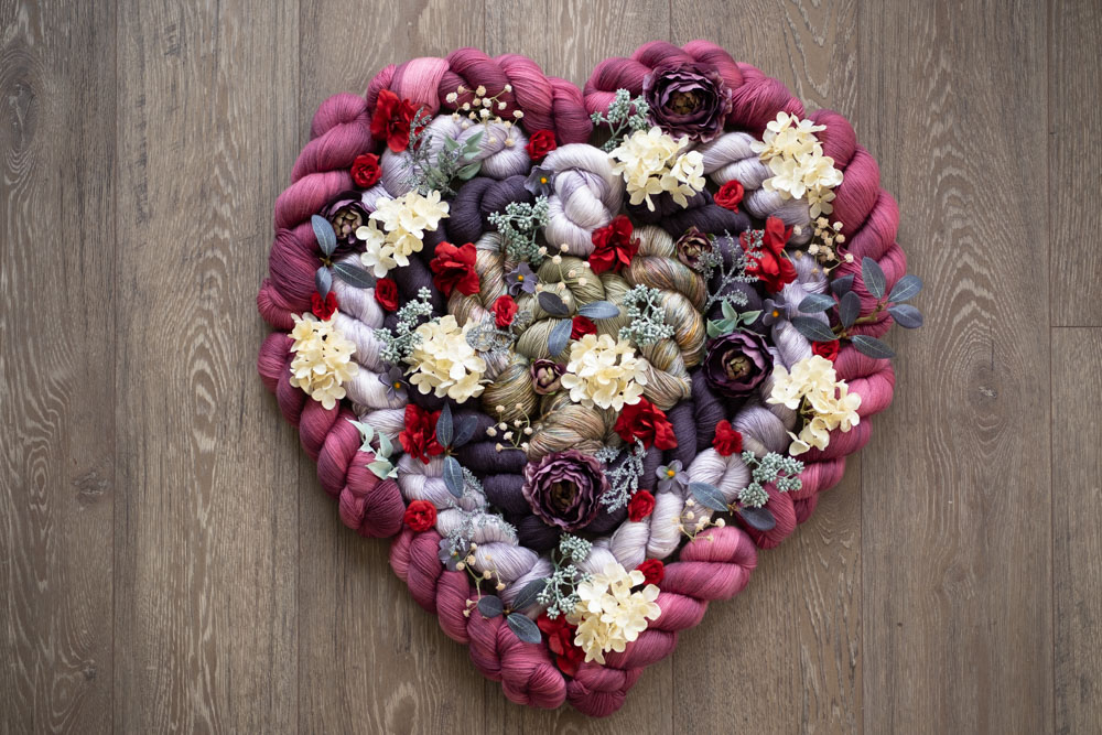 A heart-shaped pile of hand-dyed yarn and dried flowers in pink, lavender, purple and more colors